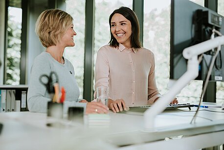 Zwei Frauen stehen an einem höhenverstellbaren Schreibtisch und lächeln sich an.